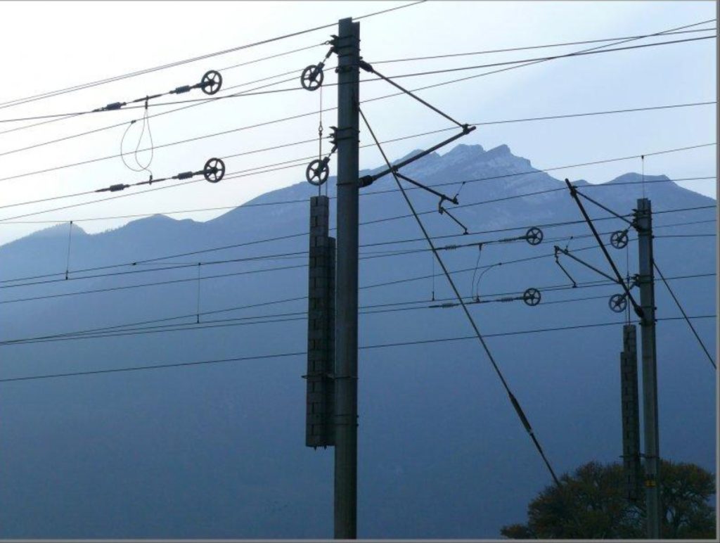 Ligne ferroviaire électrifiée au pied des montagnes, avec poteaux et câbles de caténaire